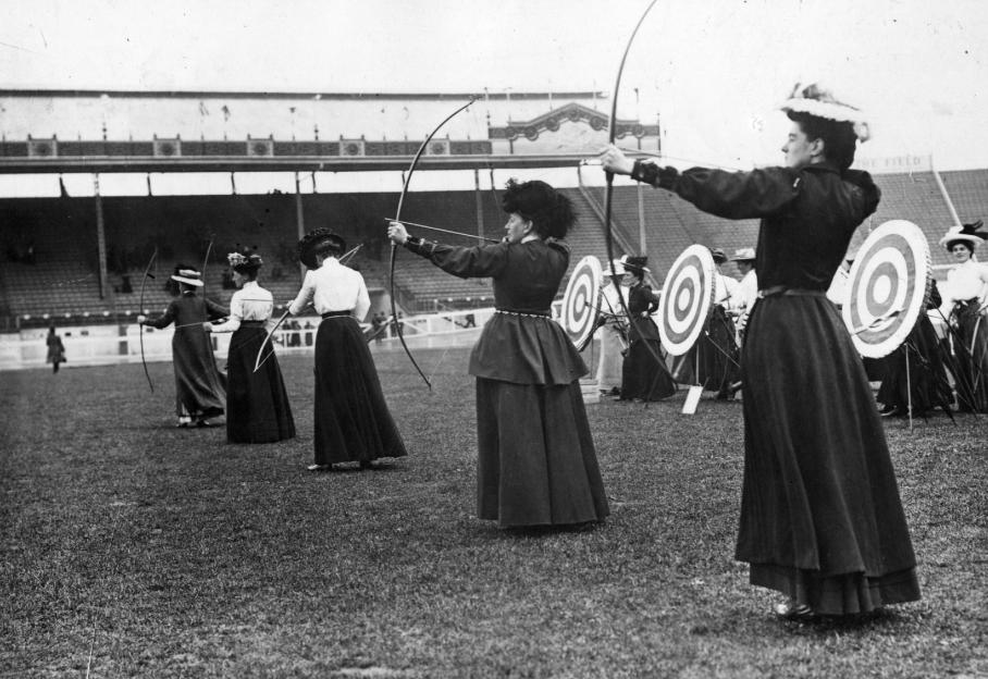 Cette image montre un groupe d'archère des années 1900, probablement pendant une compétition. Elles sont en robe. Derrière elles, on voit un gradin mais aussi des cibles. En somme, elles représentent l'histoire du tir à l'arc.